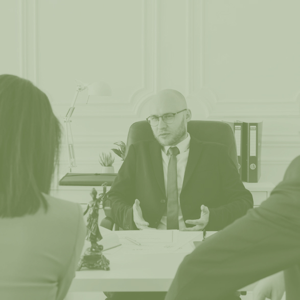 Three people in formal attire have a serious discussion in an office; a bald man in glasses sits behind a desk speaking to two others whose backs are visible, with papers and legal scales on the desk.